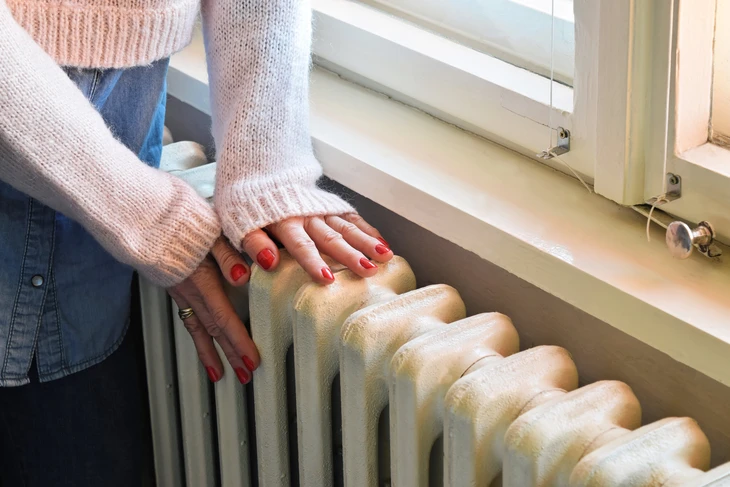 Une femme en pull rose et jeans place ses mains sur un radiateur à ailettes près d'une fenêtre pour tester la chaleur