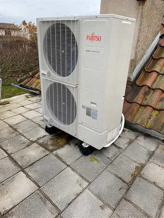 Extérieur résidentiel avec unité de climatisation Fujitsu installée sur terrasse carrelée près d'un toit en tuiles