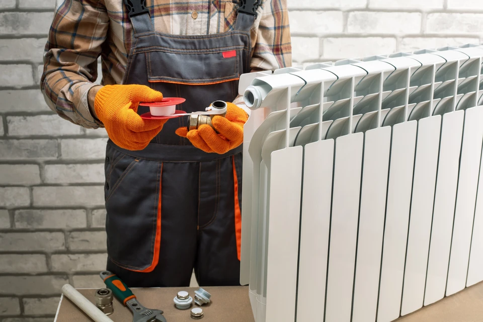 Technicien en salopette et gants orange effectue la maintenance d'un radiateur à ailettes en aluminium blanc dans un intérieur résidentiel